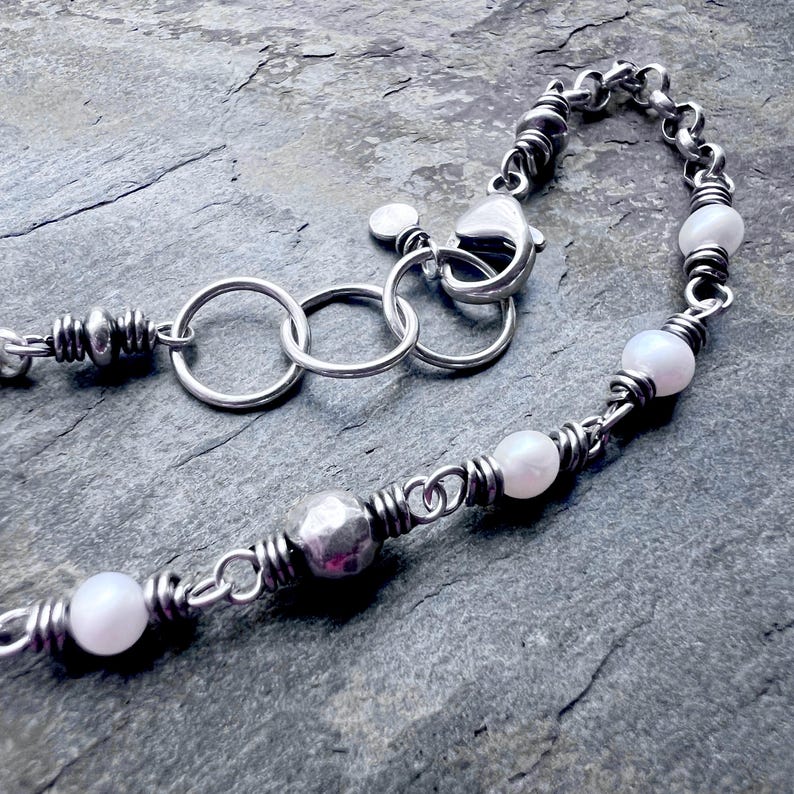 May include: A silver necklace with a chain and clasp. The necklace features white pearl beads and a hammered silver bead. The chain is made of small, twisted silver links.