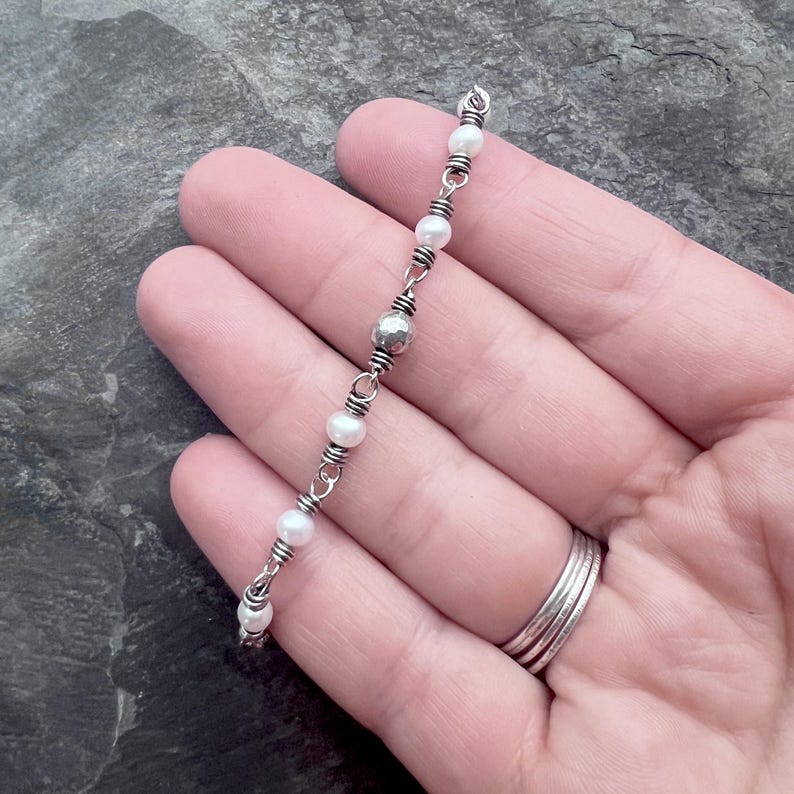 May include: Delicate silver bracelet featuring small, white pearls and a single, textured silver bead. The pearls are strung together with silver wire, creating a simple yet elegant design. The bracelet is shown on a hand.
