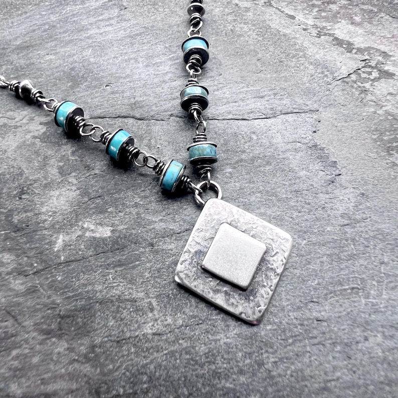 May include: Silver-toned necklace featuring turquoise-colored beads and a textured, square pendant with a smaller square inset. The necklace has a delicate chain and a handcrafted appearance, suitable for everyday wear.