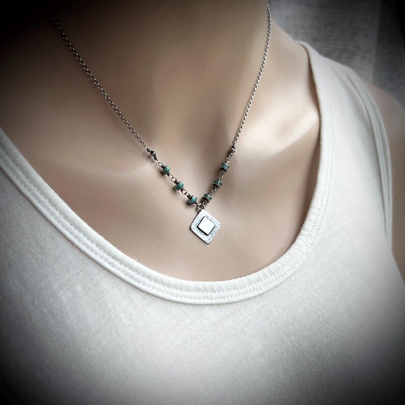 May include: A silver necklace with a diamond-shaped pendant and small turquoise beads. The necklace is worn on a person wearing a white tank top. The pendant has a textured surface and a small square in the center.