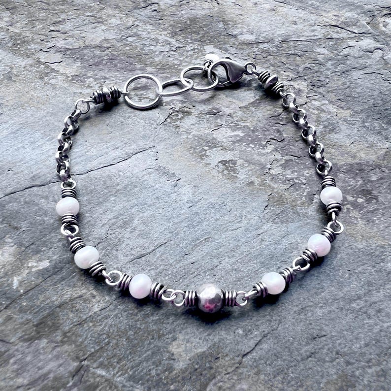 May include: A silver bracelet featuring a delicate chain design interspersed with small, round, pale pink beads. The bracelet includes a larger, textured silver bead and a secure clasp.