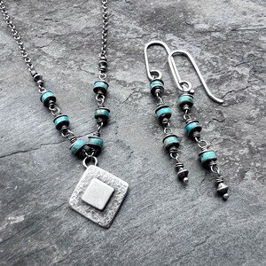 May include: A silver necklace and matching earrings set. The necklace features a diamond-shaped pendant with a square center, and turquoise beads. The earrings have a similar design, with turquoise beads and silver accents. The jewelry is displayed on a gray surface.