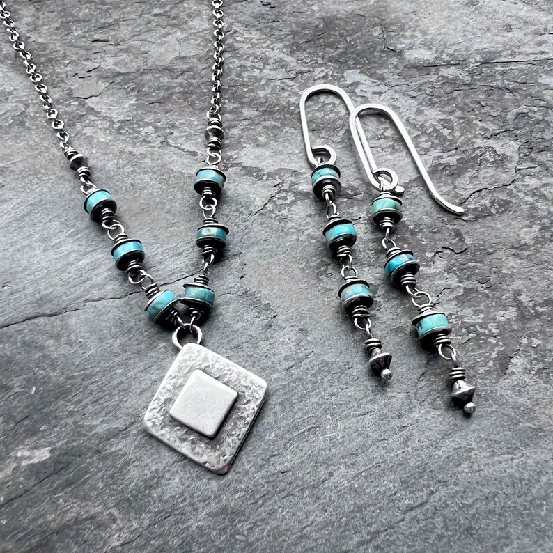 May include: A silver necklace and matching earrings set. The necklace features a diamond-shaped pendant with a square center, and turquoise beads. The earrings have a similar design, with turquoise beads and silver accents. The jewelry is displayed on a gray surface.
