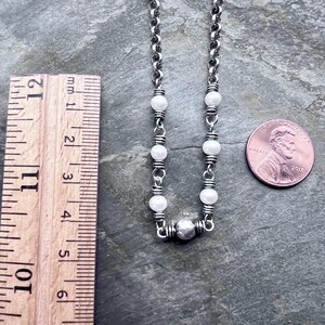 May include: A silver chain necklace with white beads and a silver pendant. The necklace is next to a ruler and a penny. The necklace is approximately 11 inches long.