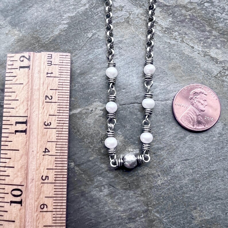 May include: A silver chain necklace with white beads and a silver pendant. The necklace is next to a ruler and a penny. The necklace is approximately 11 inches long.