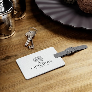 May include: White leather luggage tag with a gray strap and buckle. The tag has a logo that says "The White Lotus Resort & Spa" with a stylized lotus flower design.