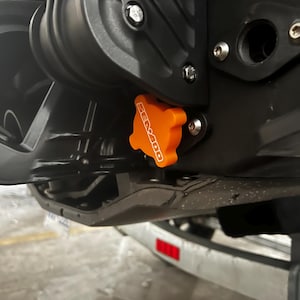 May include: A close-up shot of a black and orange Sea-Doo component. The orange part, bearing the Sea-Doo logo, appears to be a cap or cover. The surrounding structure is black, with bolts and a glimpse of a trailer in the background.