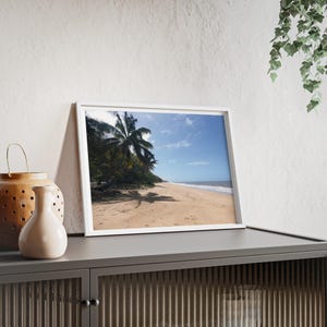 Könnte beinhalten: Ein gerahmtes Foto einer Strandszene. Das Bild zeigt einen Sandstrand, Palmen und einen blauen Himmel mit Wolken. Das Foto befindet sich in einem weißen Rahmen und wird auf einem grauen Schrank ausgestellt.