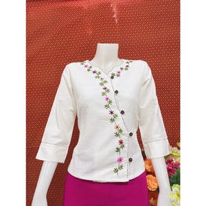 May include: White cotton blouse with a floral embroidery design on the front. The blouse has a V-neckline and a button closure on the left side.