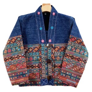 May include: A long-sleeved jacket featuring a dark blue denim upper section and a lower section adorned with a multi-colored geometric pattern. The jacket has a V-neckline with small floral embroidery, three large brown buttons, and long sleeves.
