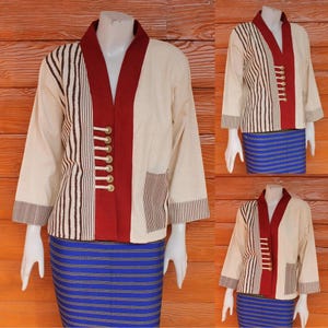 Puede incluir: Chaqueta color crema con cuello y detalles en rojo, con rayas verticales marrones en un lado y un bolsillo. La chaqueta tiene botones dorados y se combina con una falda a rayas azules. Las mangas son de tres cuartos.