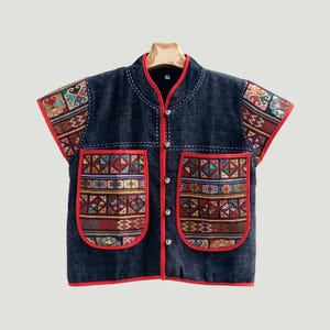 May include: A dark blue sleeveless waistcoat featuring a mandarin collar and a front button closure. The garment is accented with red trim and two large front pockets, both decorated with a colourful geometric pattern in shades of red, blue, and gold.