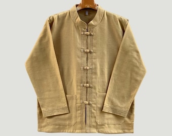 May include: A tan-colored jacket with a mandarin collar and long sleeves. The jacket features a series of decorative buttons down the front and two front pockets. The fabric has a subtle textured appearance, and the jacket is hanging on a wooden hanger.