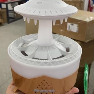 May include: A white and brown essential oil diffuser with a unique design. The top resembles a UFO, with a round base and a central column. The base is brown with control buttons. The diffuser is held in a hand.
