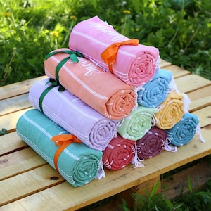 May include: A stack of rolled-up beach towels in various colors, including pink, orange, and teal, with white stripes. Each towel is tied with a ribbon. The towels are arranged on a wooden surface, with green foliage in the background.
