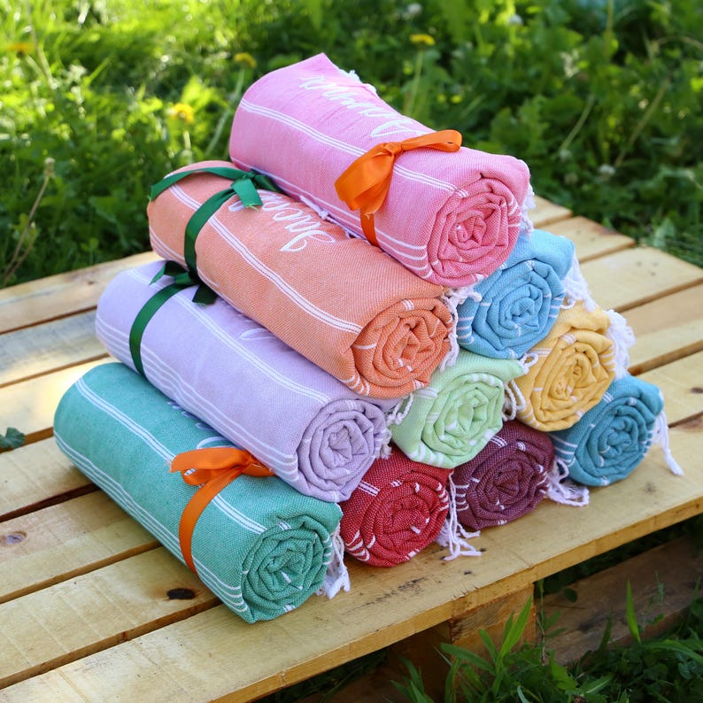 May include: A stack of rolled-up beach towels in various colors, including pink, orange, and teal, with white stripes. Each towel is tied with a ribbon. The towels are arranged on a wooden surface, with green foliage in the background.