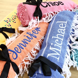 K&ouml;nnte beinhalten: Gerollte, personalisierte Strandt&uuml;cher in verschiedenen Farben, darunter Gelb, Rosa, Orange, Blau und T&uuml;rkis. Jedes Handtuch ist mit einem schwarzen Band gebunden und mit einem Namen in einer Kontrastfarbe bestickt. Die T&uuml;cher haben wei&szlig;e Fransen.
