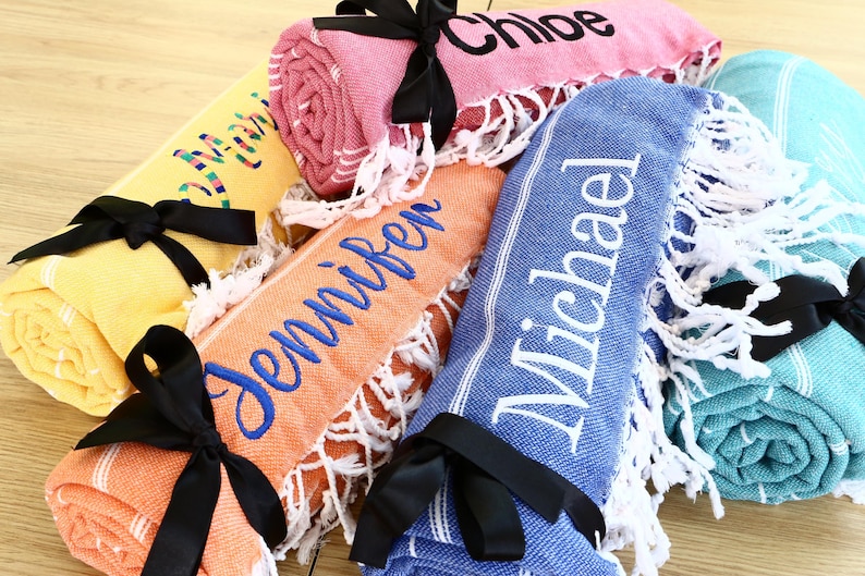 K&ouml;nnte beinhalten: Gerollte, personalisierte Strandt&uuml;cher in verschiedenen Farben, darunter Gelb, Rosa, Orange, Blau und T&uuml;rkis. Jedes Handtuch ist mit einem schwarzen Band gebunden und mit einem Namen in einer Kontrastfarbe bestickt. Die T&uuml;cher haben wei&szlig;e Fransen.
