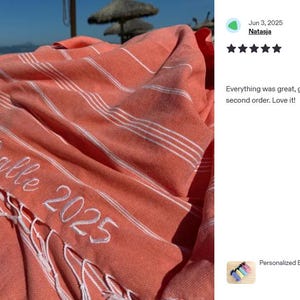 Puede incluir: Toalla de playa de color coral con rayas blancas y flecos. La toalla tiene el nombre "Malle" y el año "2025" bordados en blanco. La toalla mide 101 cm x 178 cm.