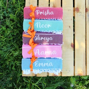 May include: A stack of five colorful personalized towels, each with a different name embroidered in white script: Prisha, Neer, Shreya, Aarna, and Emma. The towels are tied together with an orange ribbon and sit on a wooden surface.
