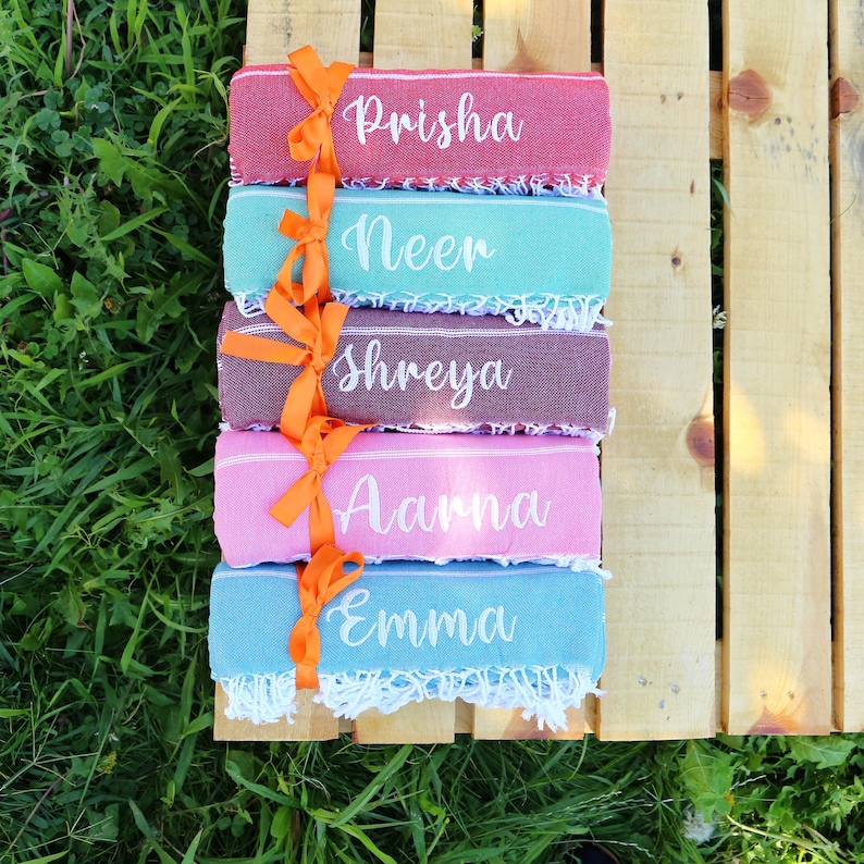 May include: A stack of five colorful personalized towels, each with a different name embroidered in white script: Prisha, Neer, Shreya, Aarna, and Emma. The towels are tied together with an orange ribbon and sit on a wooden surface.