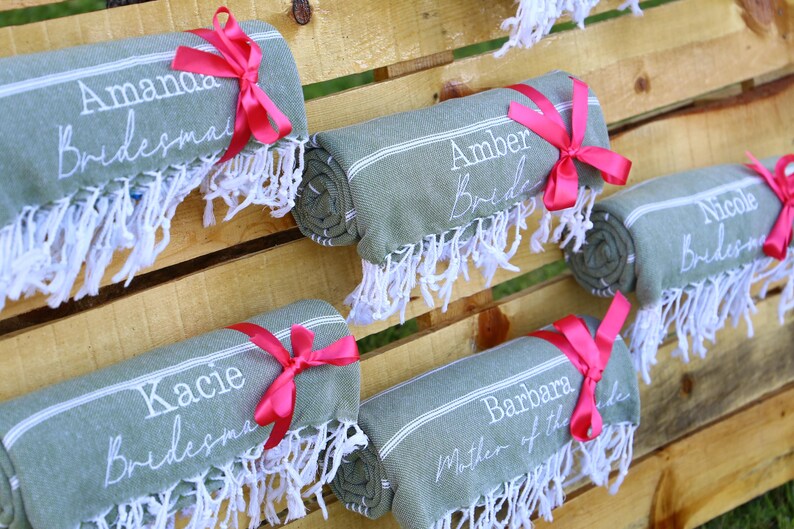 May include: Rolled green towels with white fringe and pink ribbons. Each towel is personalized with a name and title in white script, such as "Amanda Bridesmaid" and "Barbara Mother of the Bride." The towels are arranged on a wooden surface.