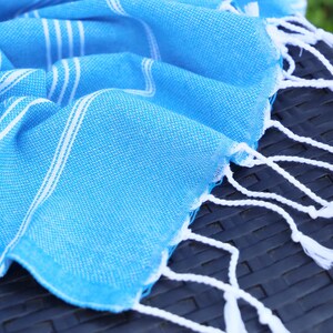 May include: A bright blue Turkish towel with white stripes and fringe. The towel is folded and draped over a dark, textured surface. The white fringe hangs down from the edge of the towel.