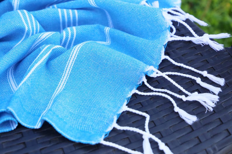 May include: A bright blue Turkish towel with white stripes and fringe. The towel is folded and draped over a dark, textured surface. The white fringe hangs down from the edge of the towel.
