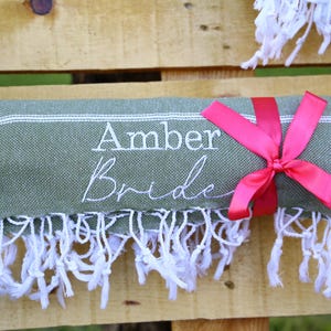 May include: A rolled green towel with white fringe and a pink ribbon. The towel has the embroidered text "Amber Bride" in white. The towel is displayed on a wooden surface.