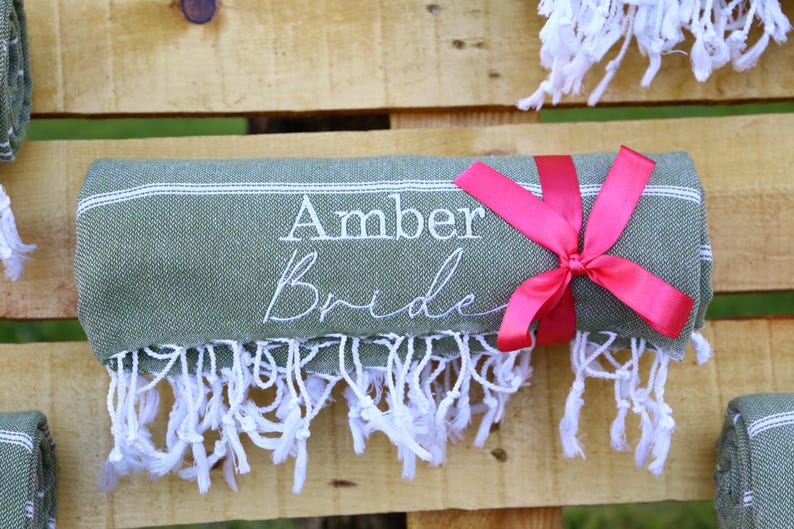 May include: A rolled green towel with white fringe and a pink ribbon. The towel has the embroidered text "Amber Bride" in white. The towel is displayed on a wooden surface.