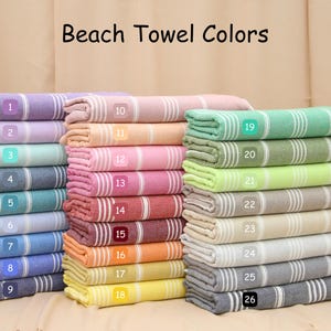 May include: Stacks of colorful beach towels in various shades, including purple, pink, green, and blue. Each towel has white stripes and a fringed edge. The text "Beach Towel Colors" is at the top.