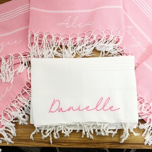 K&ouml;nnte beinhalten: Personalisierte rosa und wei&szlig;e Strandt&uuml;cher mit wei&szlig;em Fransen. Die Handt&uuml;cher haben wei&szlig;e Streifen und sind mit Namen in Schreibschrift bestickt. Ein wei&szlig;es Handtuch mit dem Namen "Danielle" ist ebenfalls abgebildet.