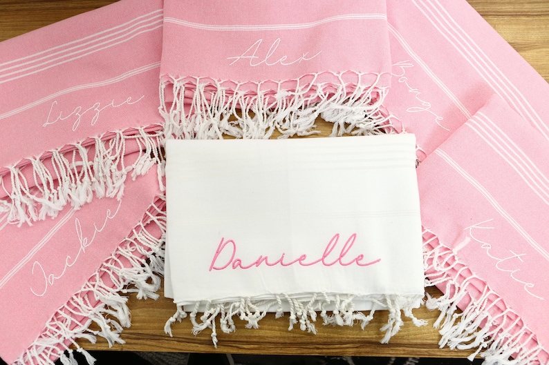 K&ouml;nnte beinhalten: Personalisierte rosa und wei&szlig;e Strandt&uuml;cher mit wei&szlig;em Fransen. Die Handt&uuml;cher haben wei&szlig;e Streifen und sind mit Namen in Schreibschrift bestickt. Ein wei&szlig;es Handtuch mit dem Namen "Danielle" ist ebenfalls abgebildet.