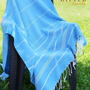 May include: A light blue towel with white stripes and fringe is draped over a black chair. The towel has the text "GIFTED towels" in the upper right corner. The towel is on a background of green grass.