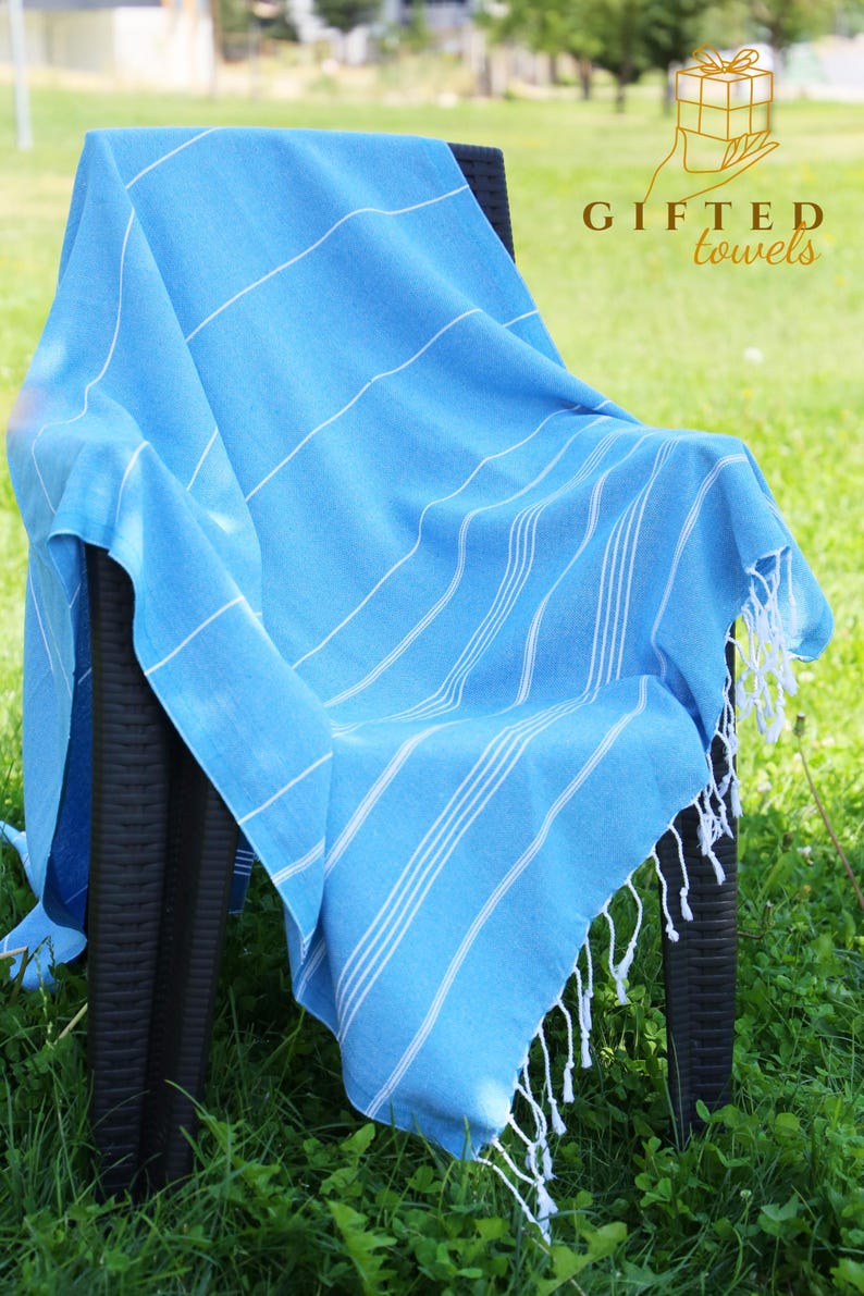 May include: A light blue towel with white stripes and fringe is draped over a black chair. The towel has the text "GIFTED towels" in the upper right corner. The towel is on a background of green grass.