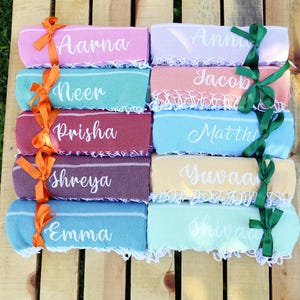 May include: A collection of personalized beach towels in various colors, each with a name in white script. The towels are tied with orange and green ribbons and arranged on a wooden surface, perfect for a summer gift.
