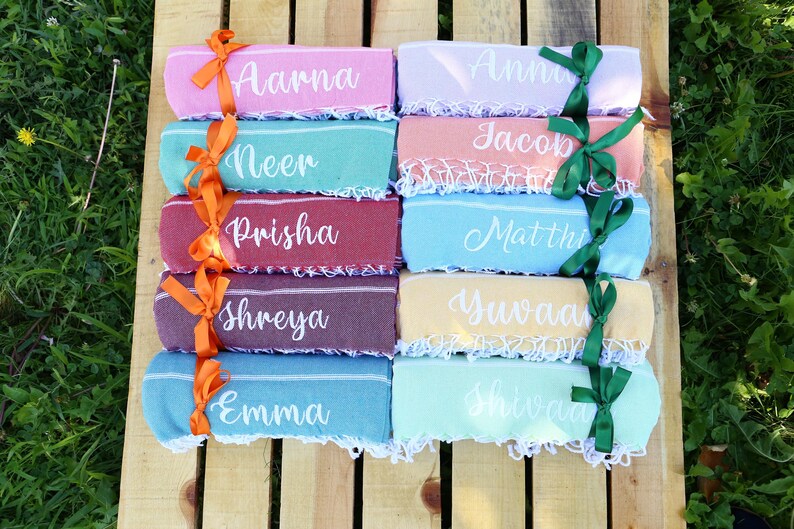 May include: A collection of personalized beach towels in various colors, each with a name in white script. The towels are tied with orange and green ribbons and arranged on a wooden surface, perfect for a summer gift.