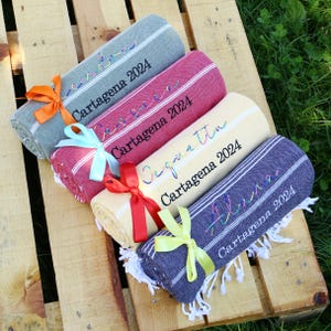 May include: Rolled towels in gray, burgundy, cream, and navy blue, each tied with a ribbon. The towels have white stripes and personalized text that reads "Cartagena 2024" and names in colorful script.
