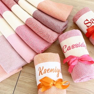 Peut inclure: Des serviettes décoratives pliées et roulées, aux tons roses, rouges et oranges, avec des motifs en losange. Certaines sont roulées et nouées avec des rubans, avec des noms comme "Sam" et "Cassidy".