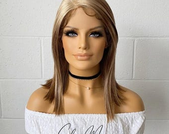 May include: A mannequin head displaying a blonde and brown wig with a blunt cut. The wig has a lace front and is styled with subtle highlights. The mannequin is wearing a black choker and a white off-the-shoulder top. The text "Glow Mane" is visible.
