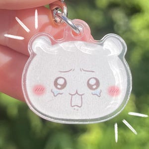 Peut inclure: Un porte-clés en acrylique transparent avec un motif en forme d'ours blanc exprimant une tristesse. L'ours a des joues roses et une petite bouche. Le porte-clés a un anneau en métal argenté et une pièce supérieure rose.