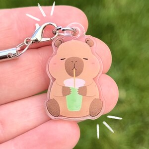 May include: A clear acrylic charm featuring a cartoon capybara drinking a green beverage. The charm has a silver clasp and a pink lanyard. The capybara is light brown with a pink blush and is holding a green drink with a white cup.