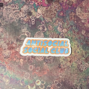 May include: A sticker with the words "ANTISOCIAL SOCIAL CLUB" in blue bubble letters with an orange outline. The sticker is on a textured background with a mix of colors including purple, brown, and blue.