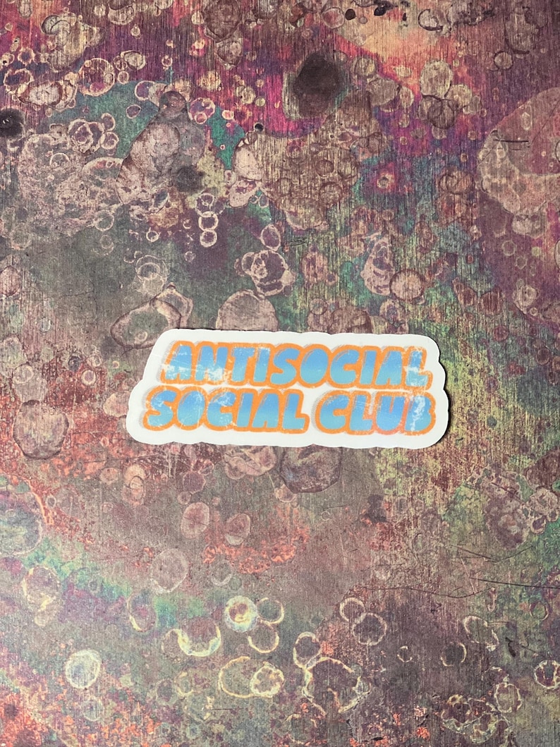 May include: A sticker with the words "ANTISOCIAL SOCIAL CLUB" in blue bubble letters with an orange outline. The sticker is on a textured background with a mix of colors including purple, brown, and blue.
