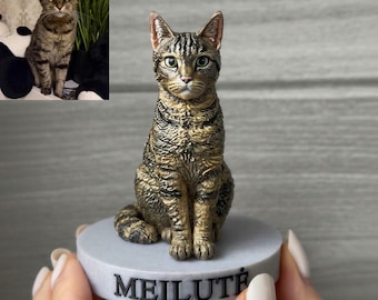 May include: A small, hand-painted figurine of a brown and black tabby cat sitting upright. The cat is mounted on a circular, light gray base with the name MEILUTĖ embossed in black capital letters on the front side of the base.
