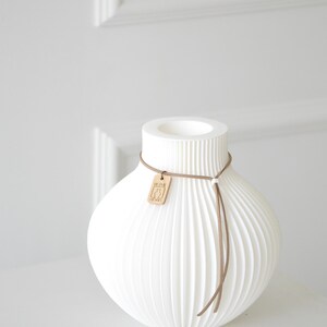 May include: White ceramic vase with a ribbed, textured design. The vase has a rounded body and a cylindrical neck. A brown leather cord with a wooden tag is wrapped around the neck. The tag has a graphic design. Home decor.