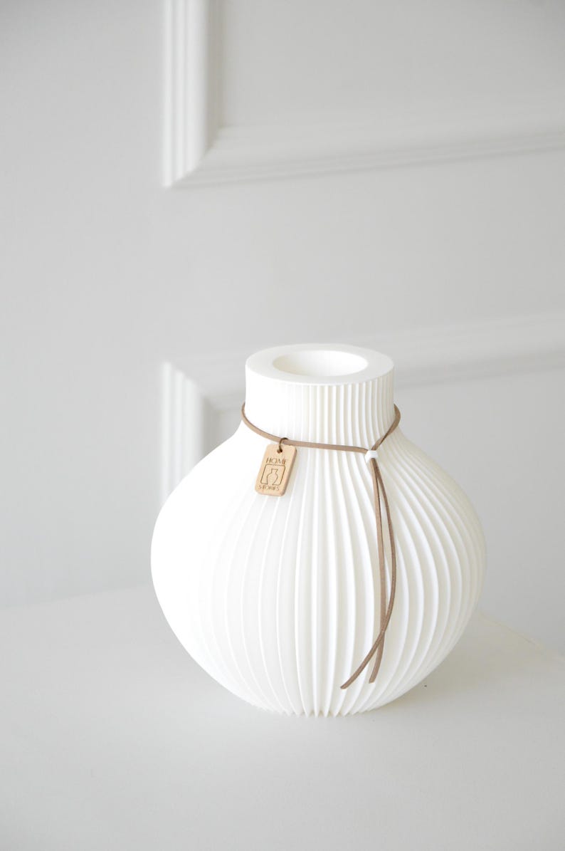May include: White ceramic vase with a ribbed, textured design. The vase has a rounded body and a cylindrical neck. A brown leather cord with a wooden tag is wrapped around the neck. The tag has a graphic design. Home decor.