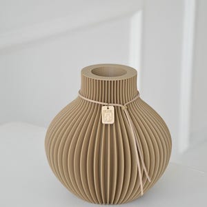 May include: A beige, ribbed vase with a round body and a narrow neck. A thin, light-colored cord wraps around the neck, holding a small wooden tag. The tag has a logo and the text "HOME STORIES".