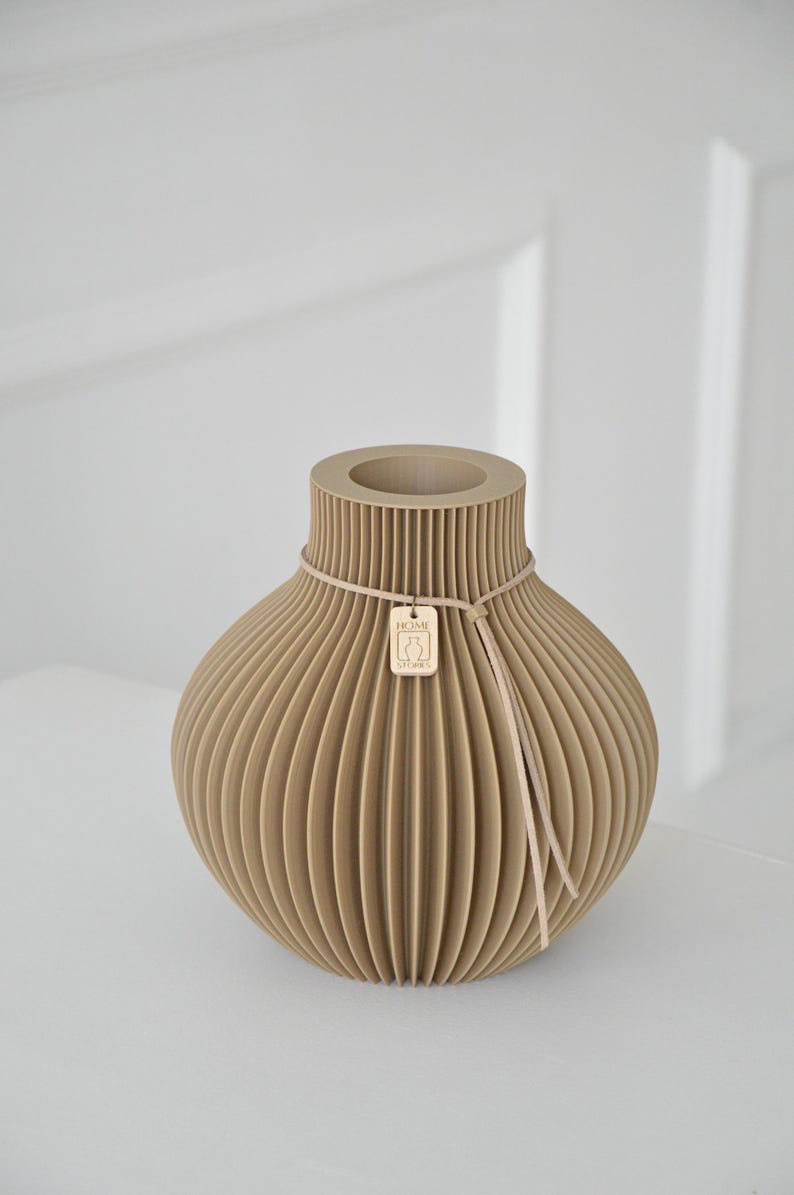 May include: A beige, ribbed vase with a round body and a narrow neck. A thin, light-colored cord wraps around the neck, holding a small wooden tag. The tag has a logo and the text "HOME STORIES".