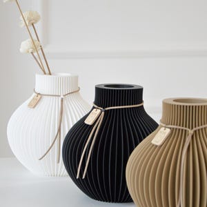 May include: Three decorative vases in white, black, and tan, each with a unique ribbed design. The white vase holds dried flowers. Each vase has a tag and is tied with a thin, light-colored cord. The vases are on a white surface.